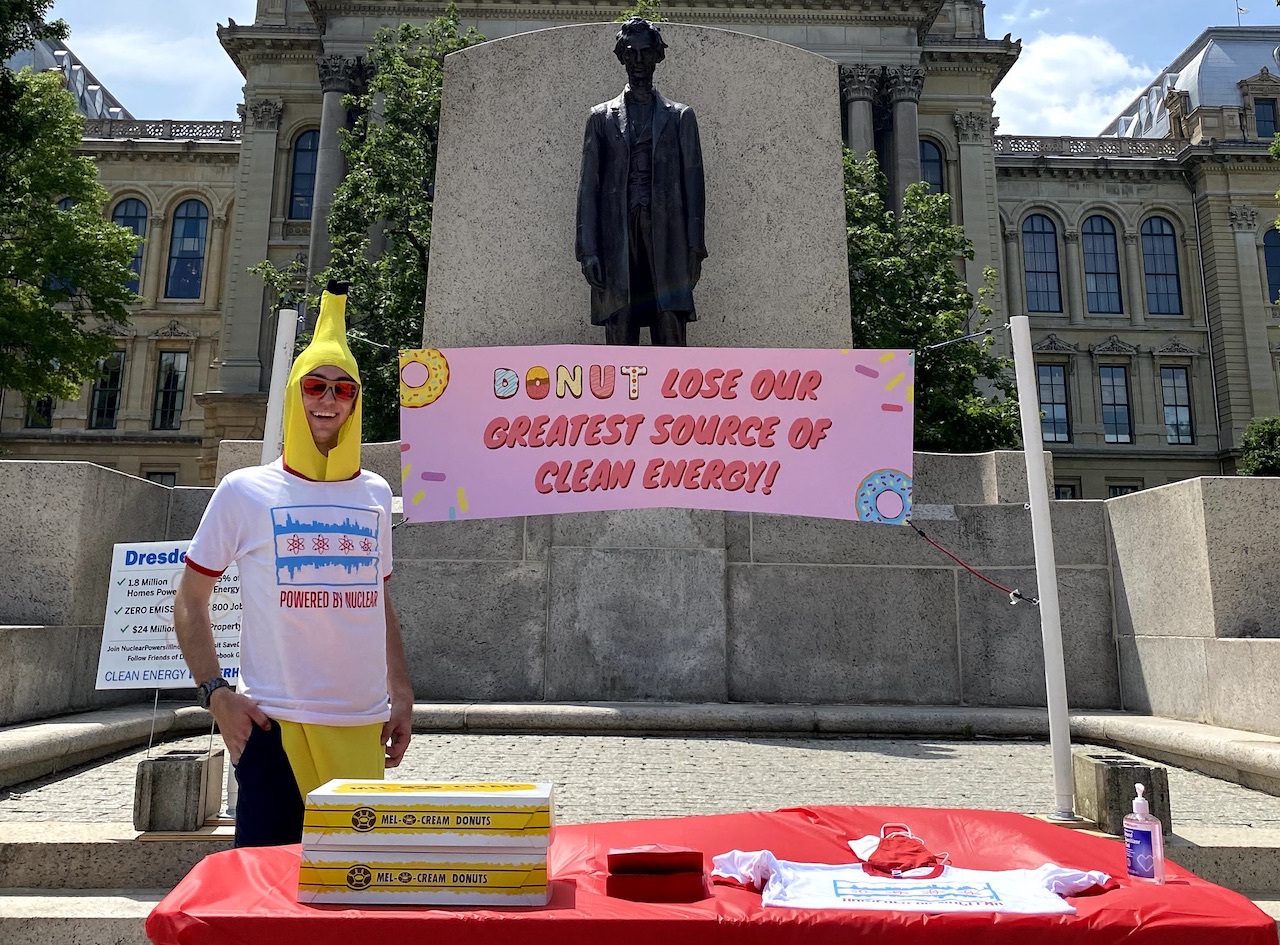 Nathan Ryan dressed in a banana suit with a shirt over it that says 'Powered by Nuclear' below a version of the Chicago flag. On the table in front of them ther are boxes of doughnuts, hand sanitizer, a shirt, and napkins. Behind them, there's a sign that says 'Donut lose our greatest source of clean energy,' and behind that is a statue of Abraham Lincoln in front of the Illinois State capitol building.