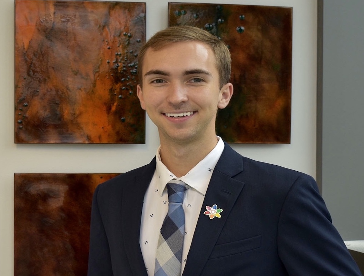 Nathan Ryan standing in front of some tiles in a blazer with a rainbow atom pin.