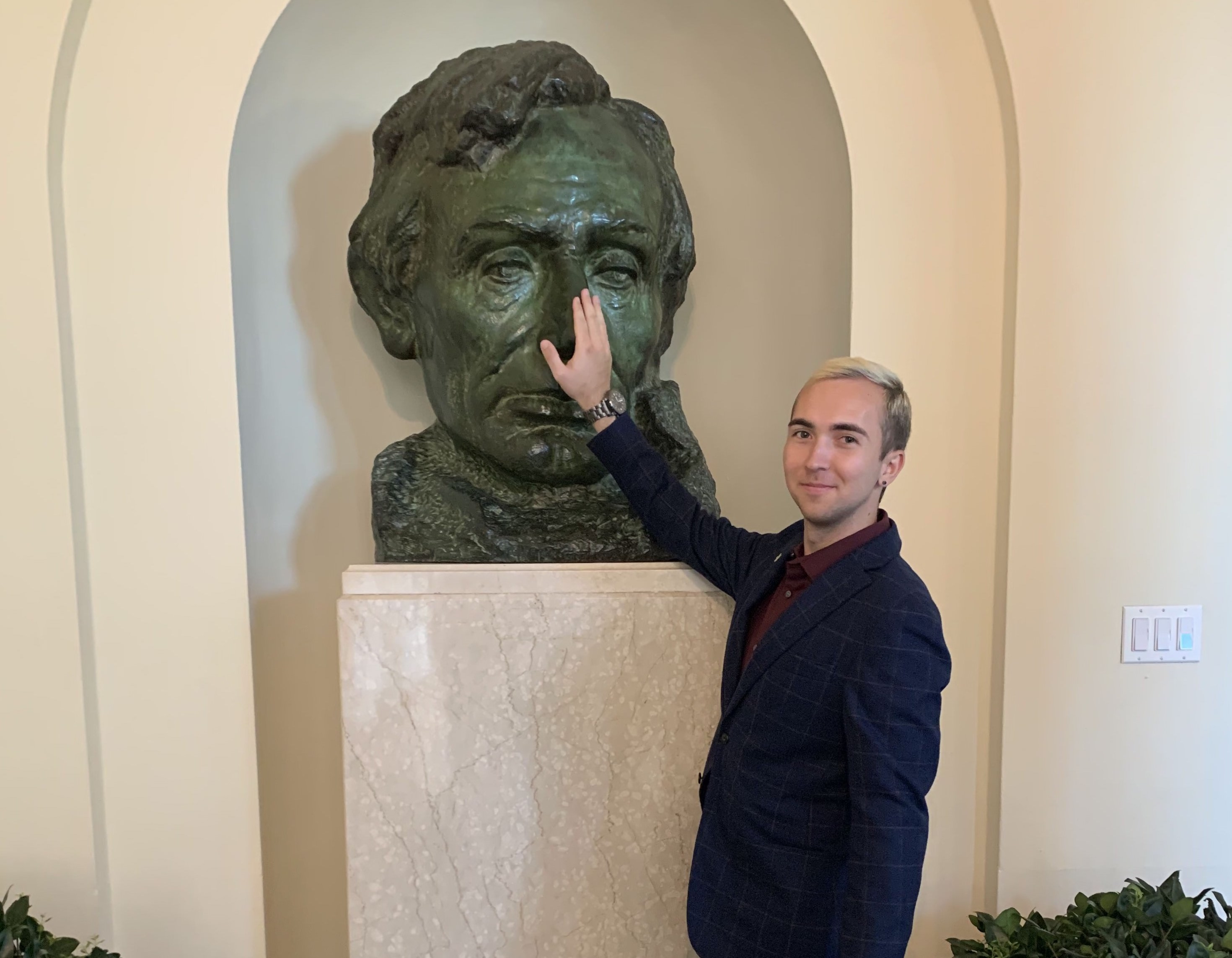 Nathan, dressed in a suit, standing in front of a bust of Lincoln touching his nose.